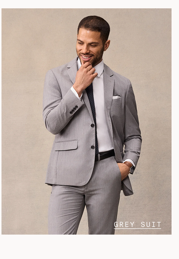 Smiling man with short dark hair touching his chin, wearing a light grey suit jacket over a white dress shirt with a navy patterned tie, white pocket square, and wristwatch, hand in pocket. GREY SUIT