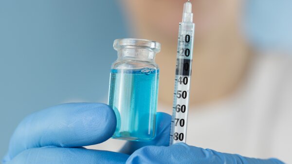 Close-up of hands wearing blue medical gloves holding a liquid blue solution in a small bottle and a syringe needle.