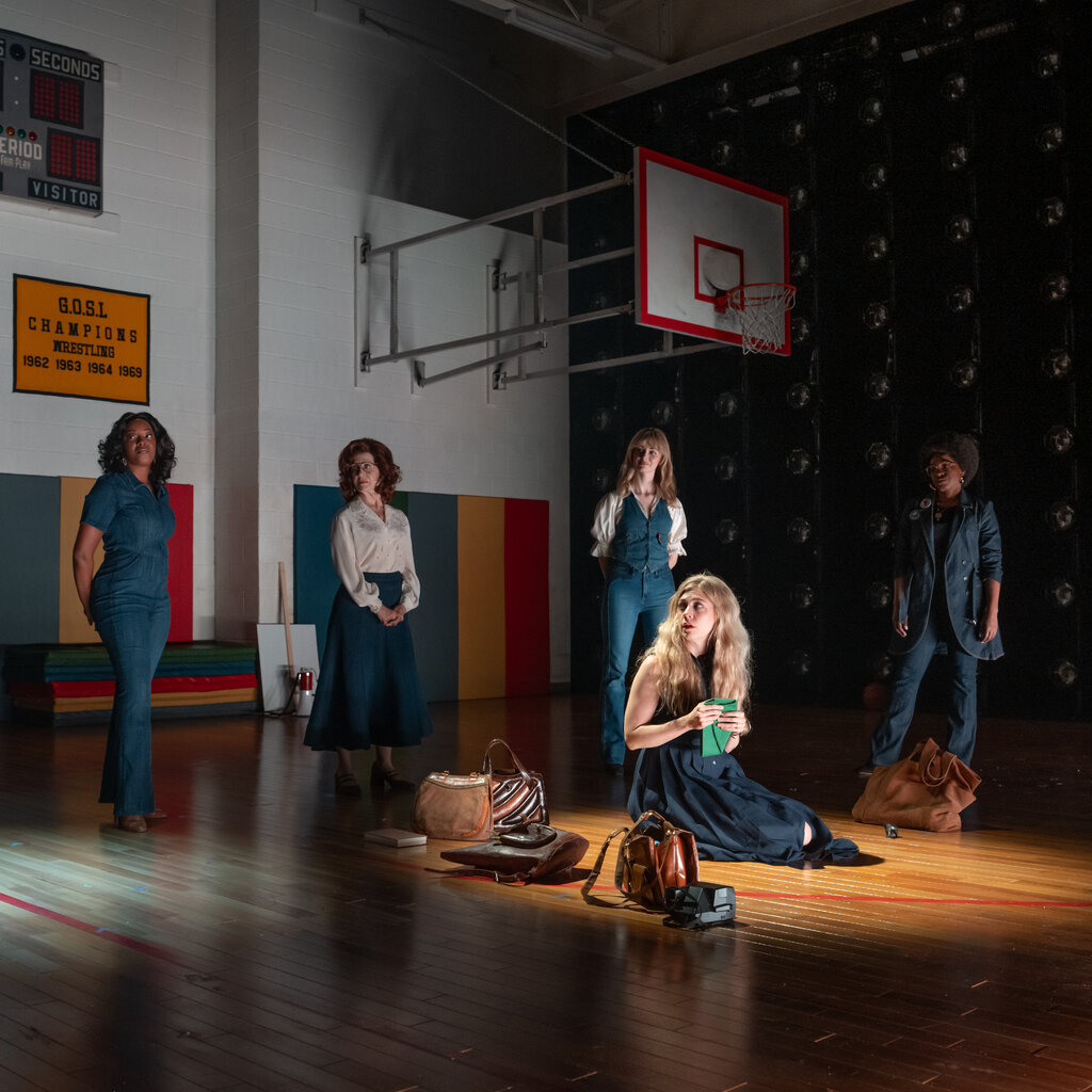 A woman is hunched on the floor of a school gym, surrounded by standing women.