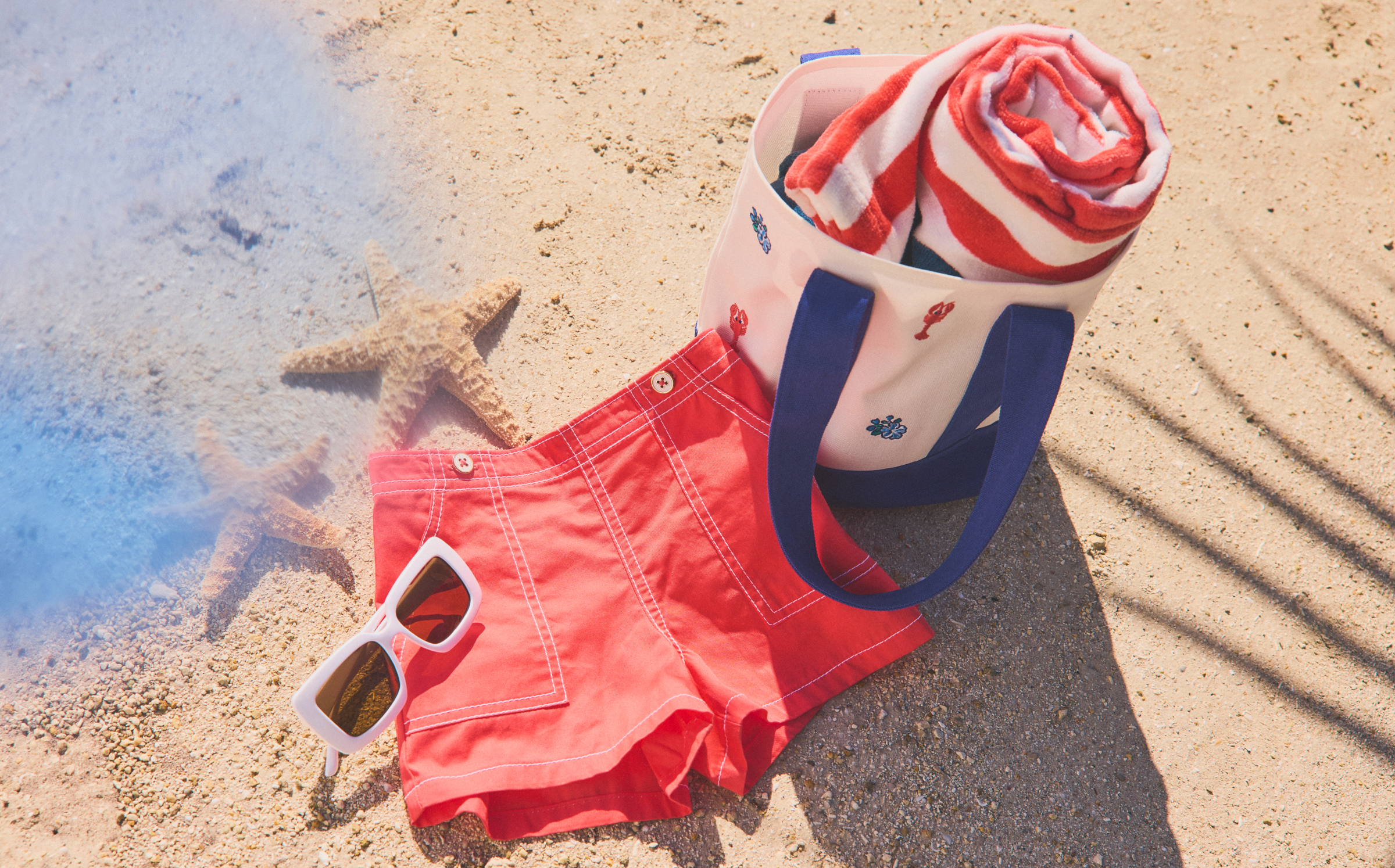 Red shorts, white tote bag with navy handles, striped beach towel, white sunglasses, and a starfish on sandy beach