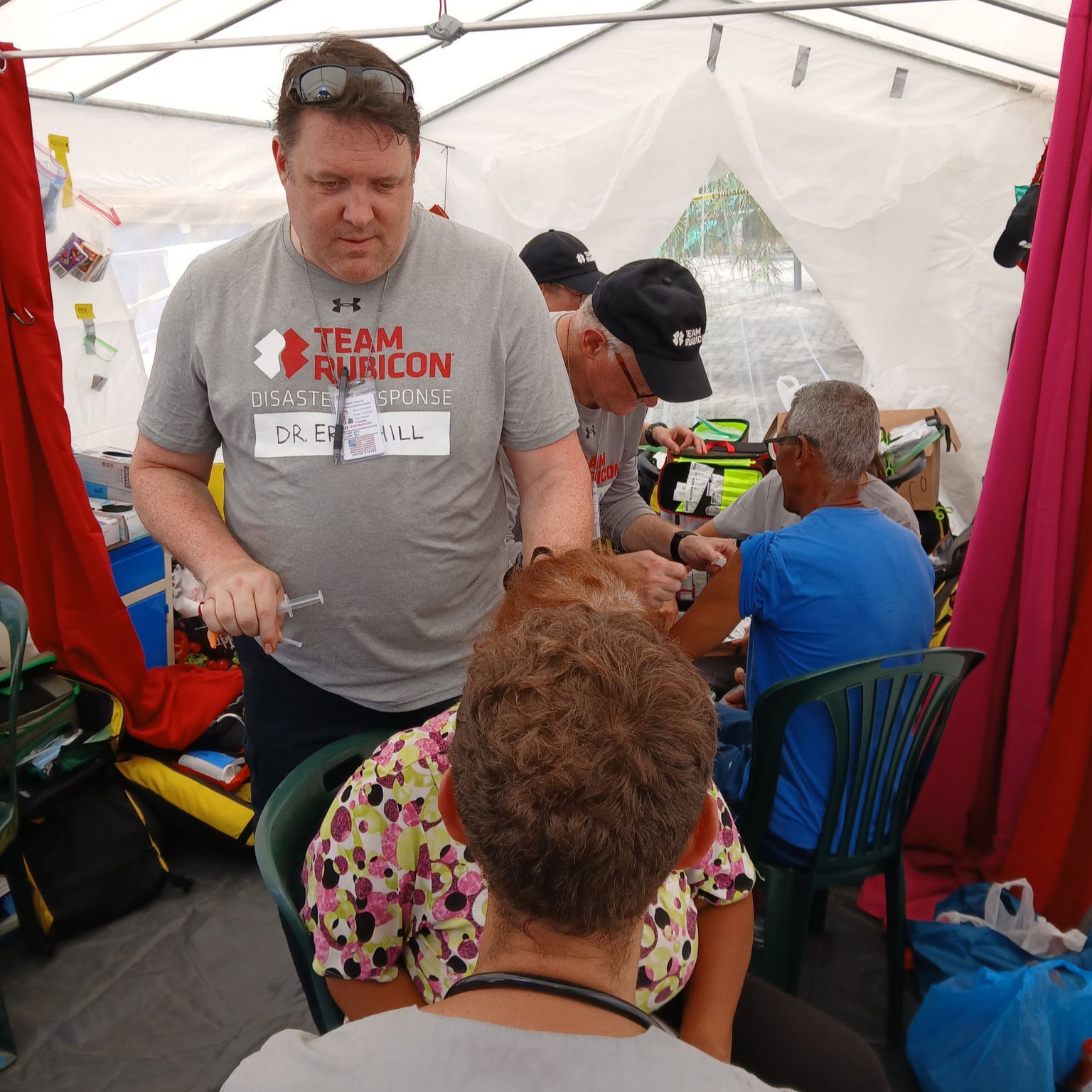 Dr. Eric Hill, a Team Rubicon medical Greyshirt, prepares a syringe while treating a patient inside a medical tent in Venezuela, as other Greyshirts and patients are cared for in the background.