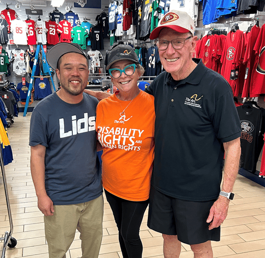 Two people wearing The Arc branded apparel, including an orange "Disability Rights Are Human Rights" T-shirt, pose with a Lids employee inside a Lids store.