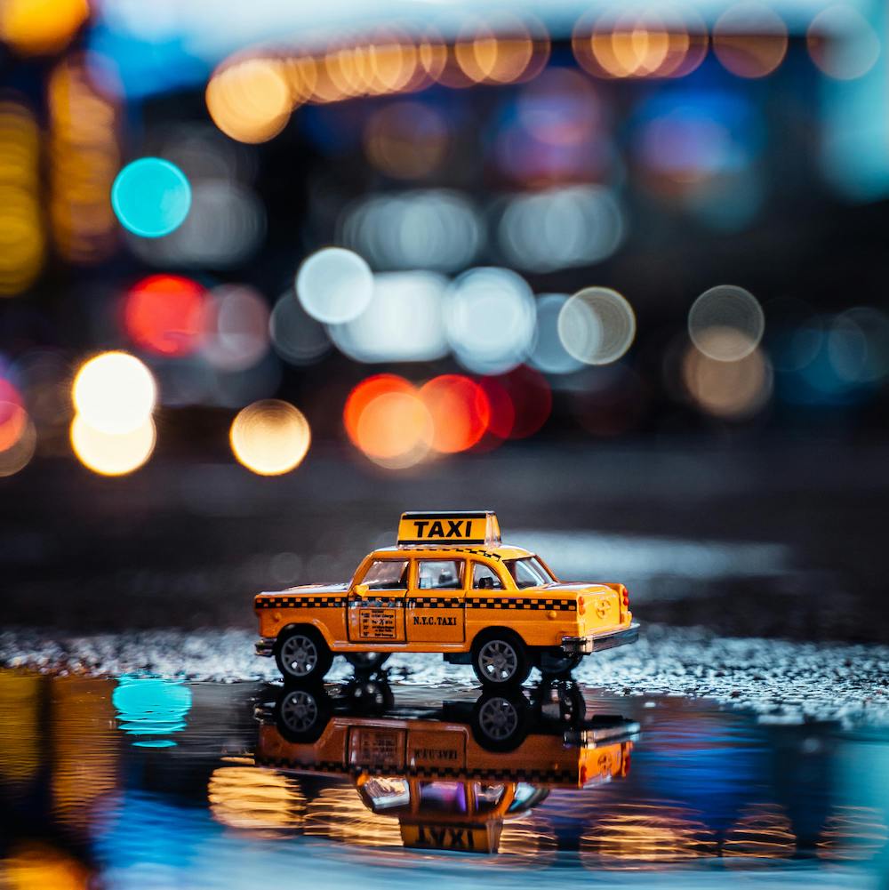 A toy taxi on a rainy city street at night. In the background are blurred city lights.