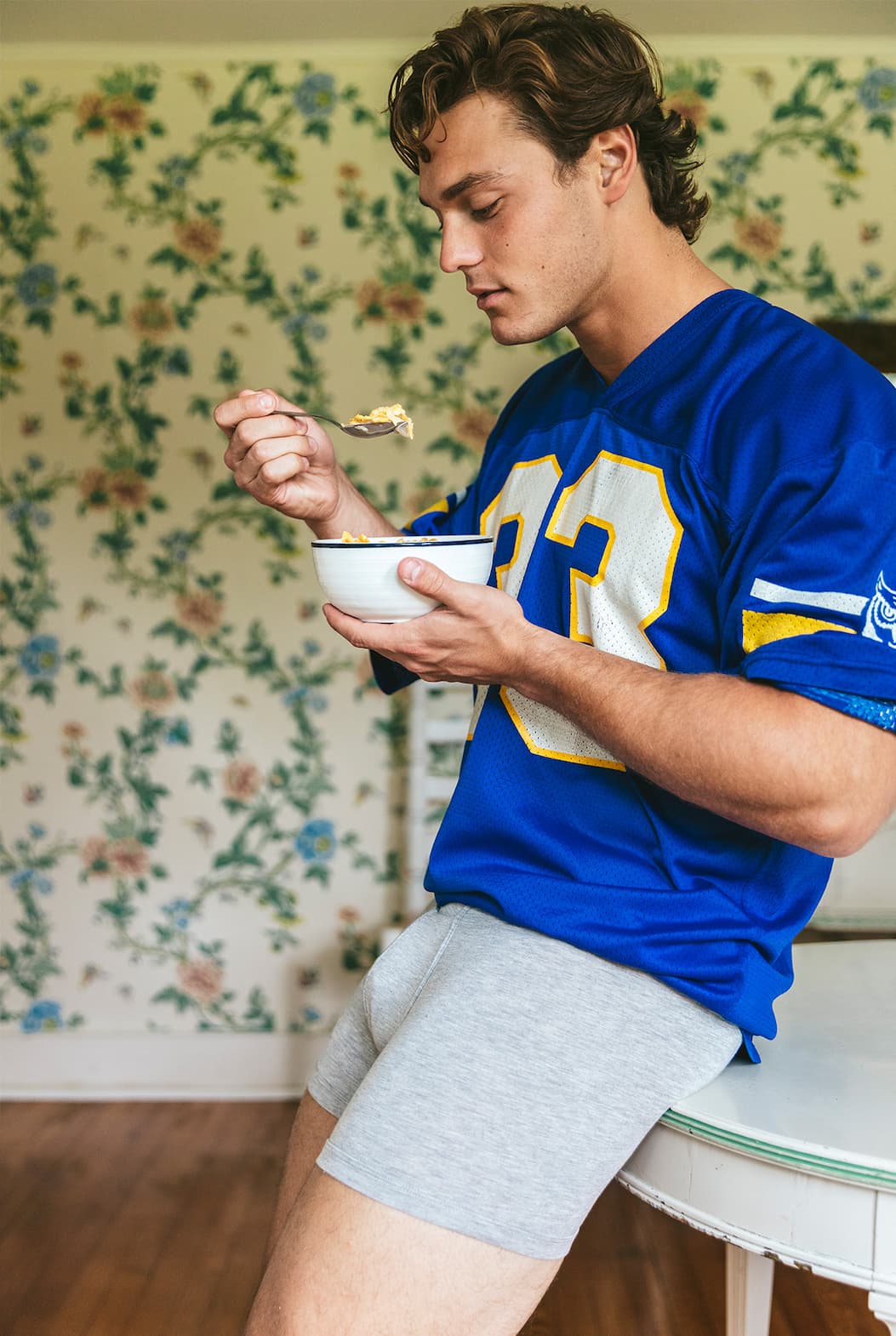 Man sitting on table eating out of a bowl and he is wearing a blue jersey and grey boxer briefs