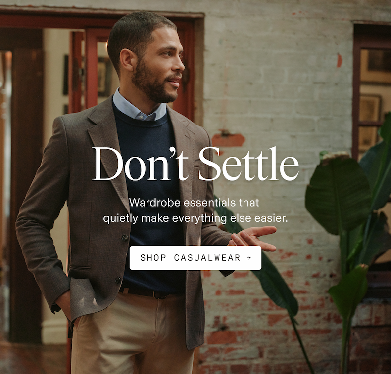 Lifestyle photograph of a man in an indoor setting with exposed brick walls and potted plant visible, wearing a brown wool blazer, navy crew neck sweater, light blue dress shirt with visible collar, and tan chinos, looking to the side. Don't Settle Wardrobe essentials that quietly make everything else easier. SHOP CASUALWEAR 