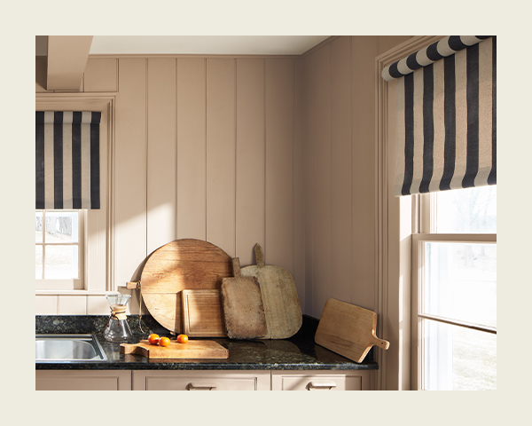 Earthy tan shiplap wall behind a kitchen counter with cutting boards, and a window with vertical striped curtains.