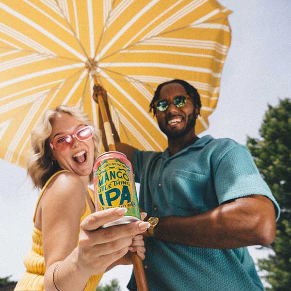 A young woman and young man smiling under a pool umbrella and holding a can of Mango Little Thing