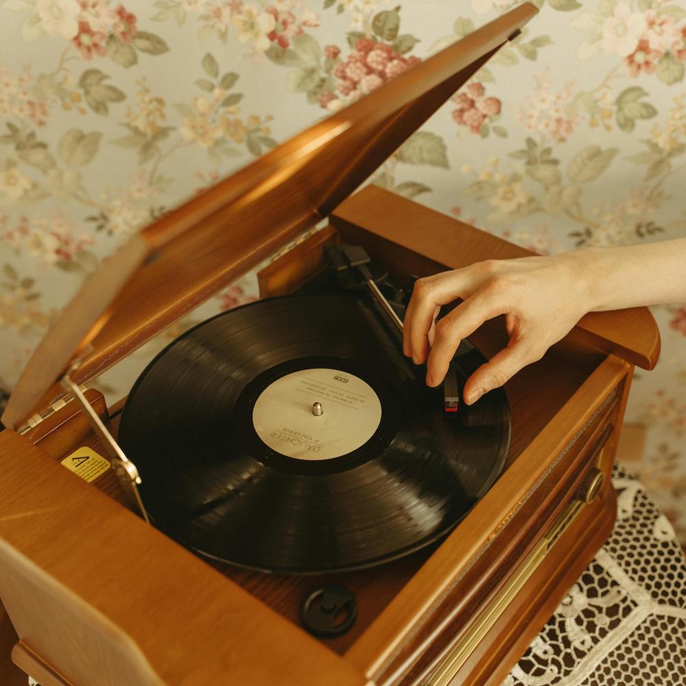 A hand places the needle on a record in a wooden turntable, located in front of floral wallpaper.
