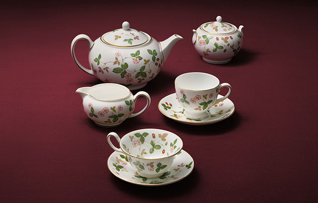 A selection of five Wild Strawberry teaware pieces presented on a red studio background. The selection features a teapot, cream jug, two teacups and saucers, and a sugar bowl.