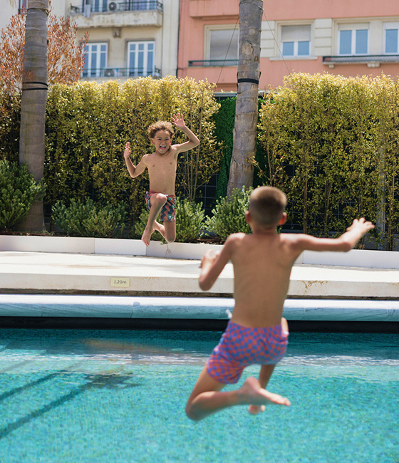 Children jump into an outdoor pool with hedges and city buildings behind.