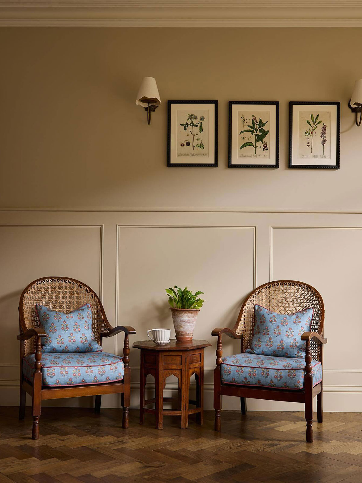 Two cane chairs with comfy looking blue silk cushions invite you to stop for a moment, enjoy a cup coffee, and relax. The pared back taupe walls give a gentle glow to the surroundings. The small potted plant on the table in its aged terracotta pot reflects the theme of the three botanical prints between the two small lamps on the wall.