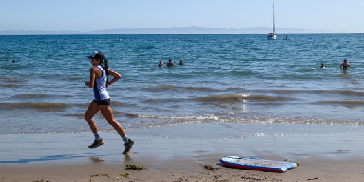 A woman jogs on a beach while people swim in the ocean along the Juan Bautista de Anza National Historic Trail