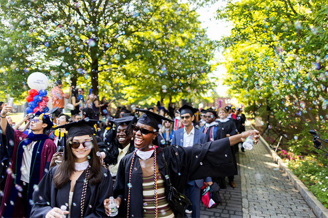 Students in graduation robes walk down Locust Walk, surrounded by bubbles.
