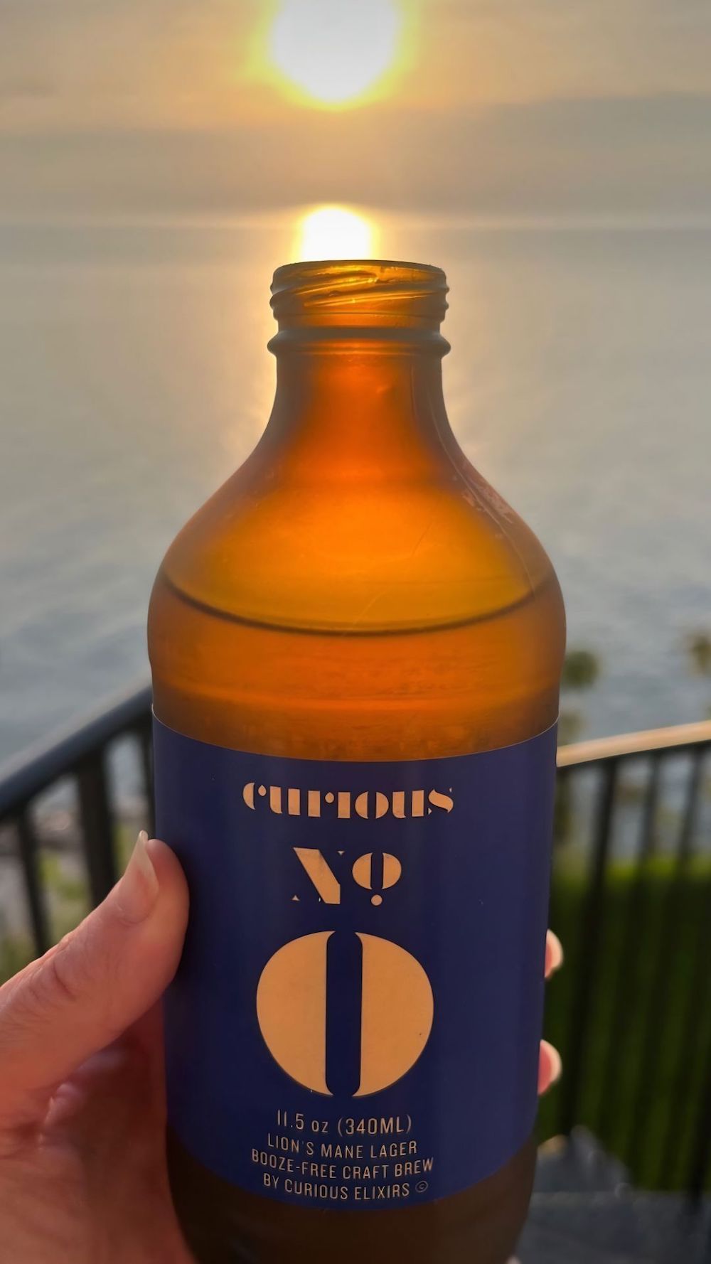 Someone holds up a bottle of Curious Zero in front of a lake, so the setting sun shines through it. 