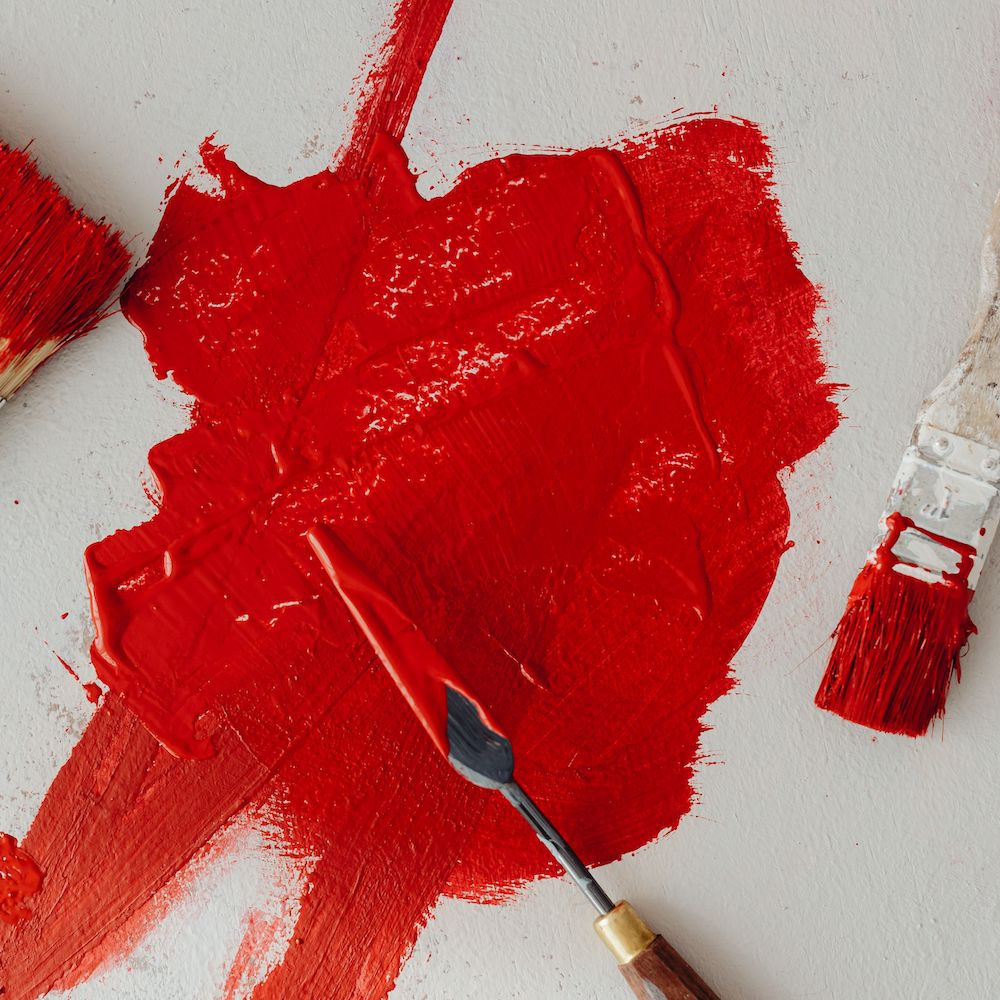 A paint spatula smears bright red paint on a white surface. 