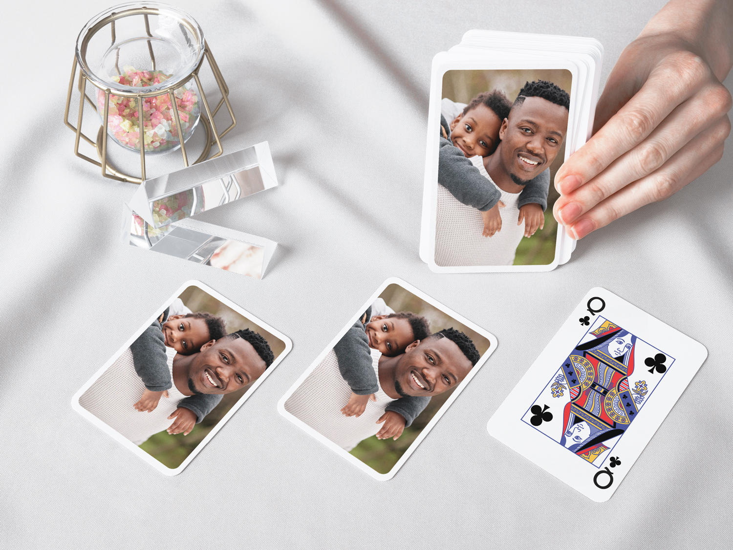 Custom playing cards featuring a smiling father and child portrait spread across a table, with a hand holding the deck beside decorative glass and mirrored accents.