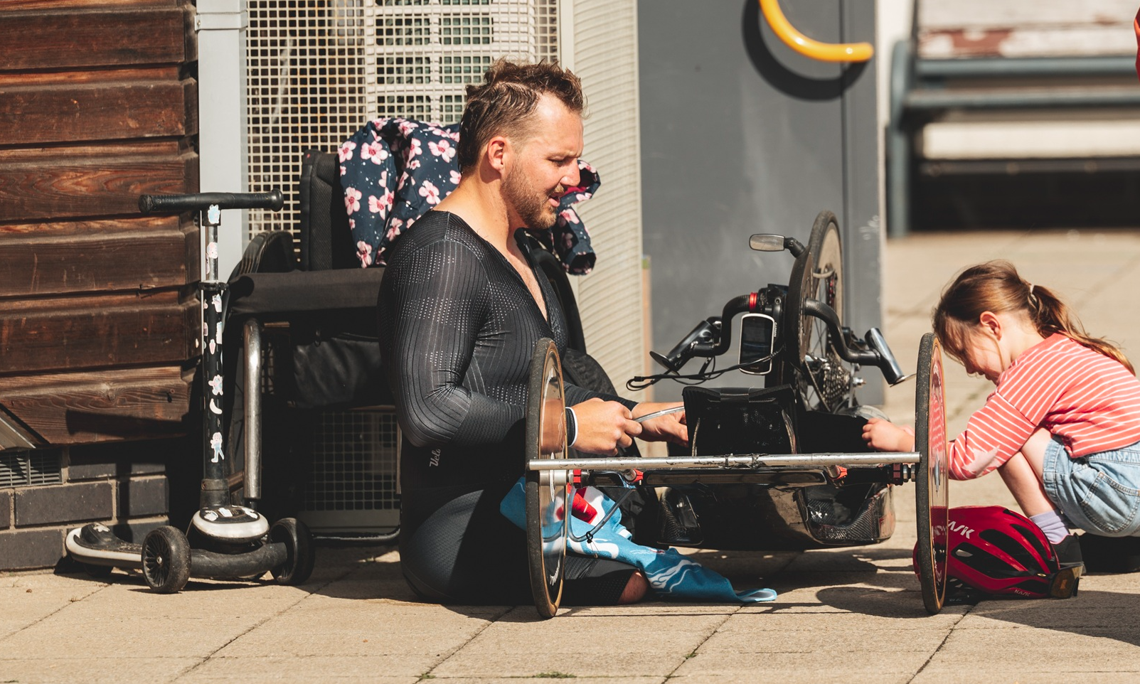 A handcyclist sits on the floor next to his bike, while a young girl crouches on the other side, looking at the bike. Her scooter is to the left of the picture.