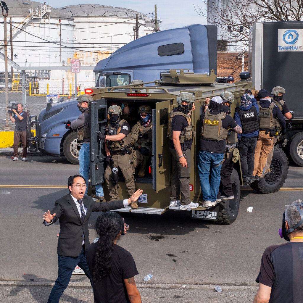 A person in a dark suit with an open mouth and outstretched arms stands on a street with debris. An olive green armored vehicle holds people in tactical gear.