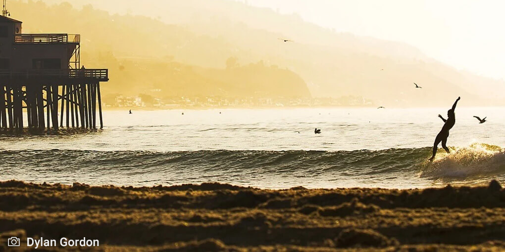 A surfer hangs five on a wave at Malibu at dawn, with the pier in the background.