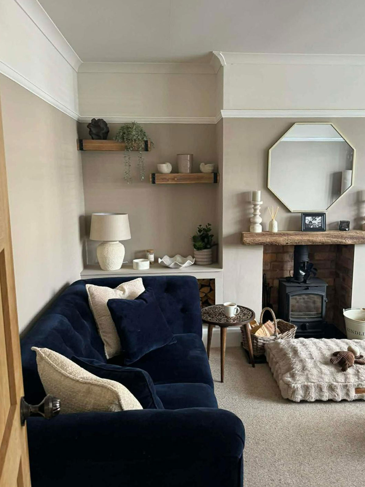 The door opens onto a cosy looking room, complete with log burner waiting to be lit. The navy velvet sofa is deep and soft, ready to sink into. A beige velvet floor cushion is perfect for curling up in front of the fire. The room is in soothing shades of cream and ecru, offset with verdant green plants trailing down from the shelves.