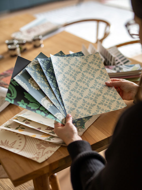 A person at a wooden table fans out patterned wallpaper samples in various colours and designs.