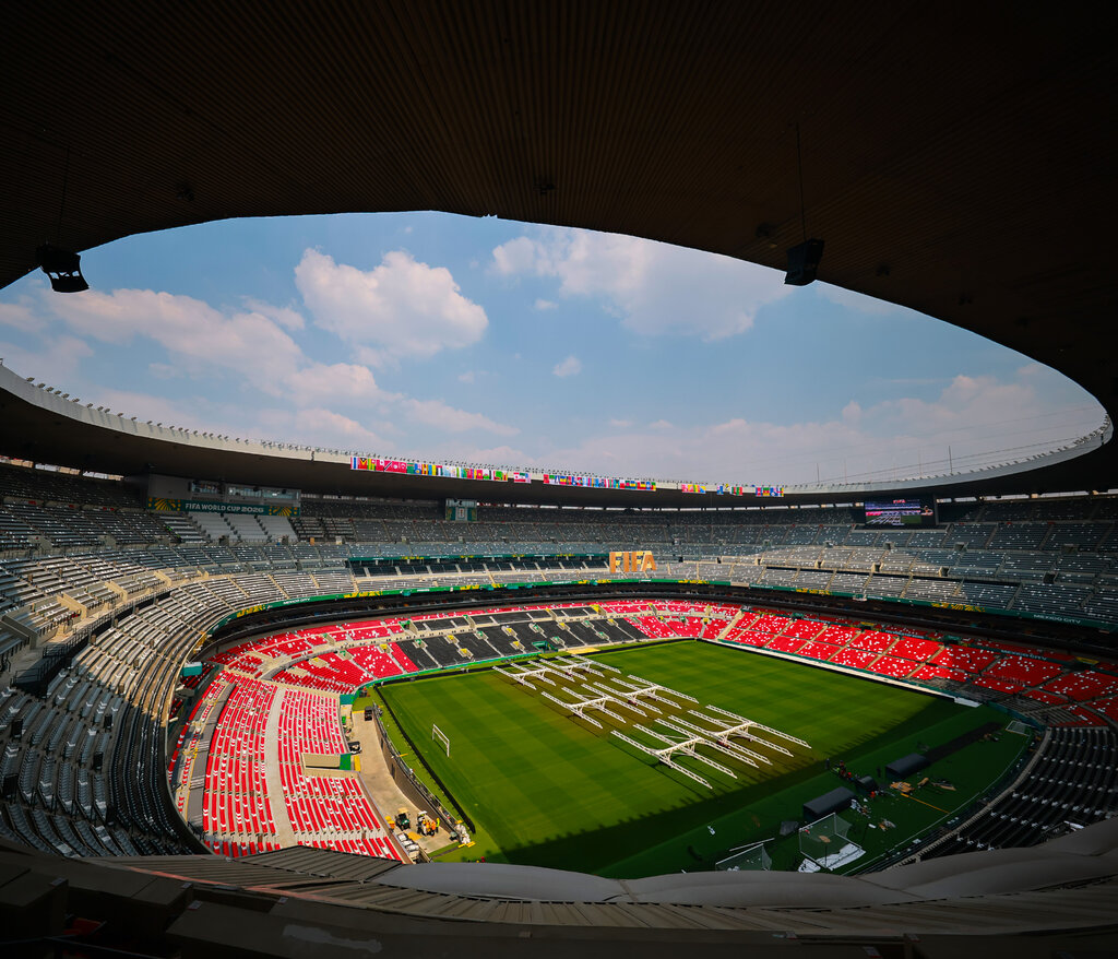 An expansive soccer stadium with tiered red, white and gray seating rows under a wide, curved roof opening that reveals a partly cloudy blue sky.