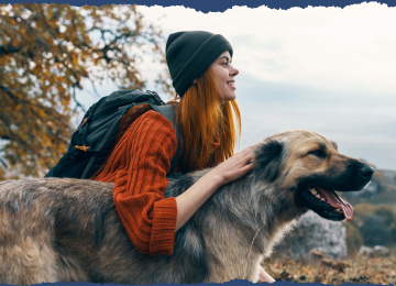 A hiker, wearing a beanie and a backpack, and her fluffy medium-sized dog are enjoying a picturesque view of rolling hills.