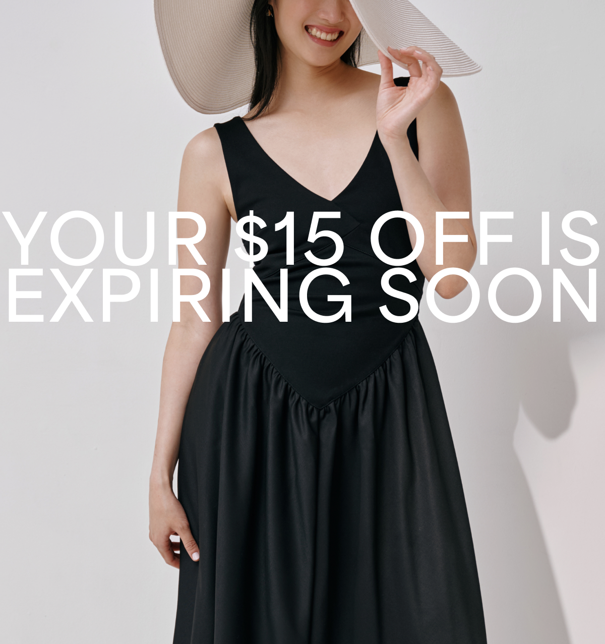 Xun is smiling in a black sleeveless Cammi Dress playfully tilts a wide-brimmed sun hat over her face. The background is minimalist and light-colored. White overlay text reads 'YOUR $15 OFF IS EXPIRING SOON'.