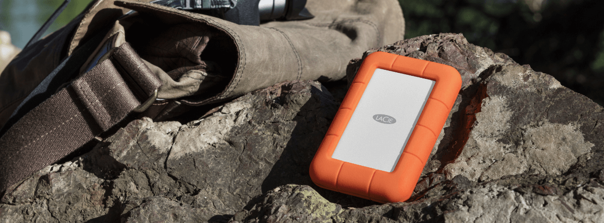 A LaCie external hard drive rests on a rock beside a brown backpack and a camera.