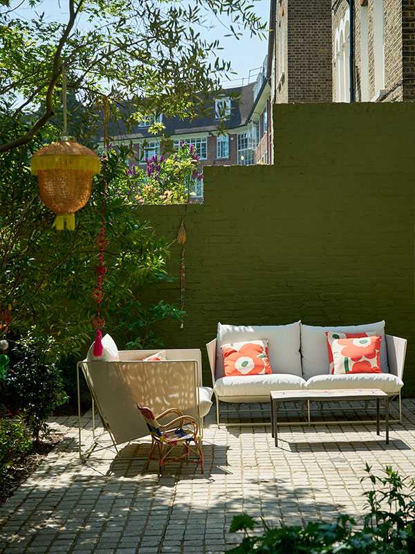 A sunlit patio with a cushioned sofa and armchair, both with bold floral cushions, surrounded by greenery and hanging woven decorations.