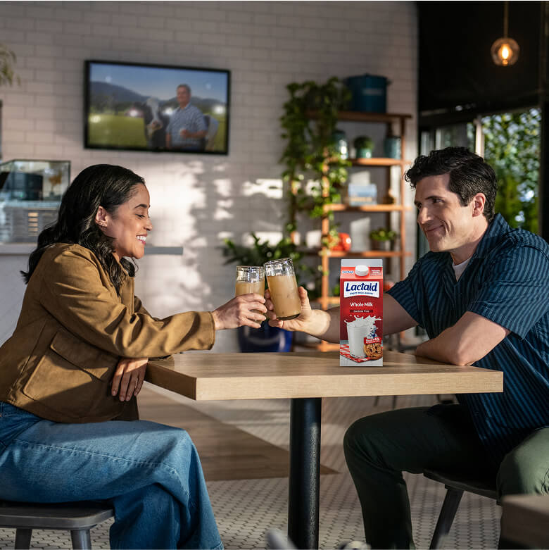 A woman on the left and a man on the right sitting at a table drinking coffee with a carton of Lactaid milk on the table.
