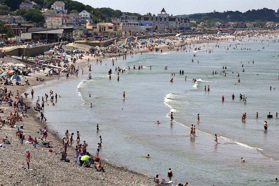 Wide photo of visitors crowding onto Nantasket Beach in Hull, MA. People are seen sitting on the small, partially rocky shore at left, while others are in the shallow water at right. Houses and buildings
lining the street along the beach are seen in the background.