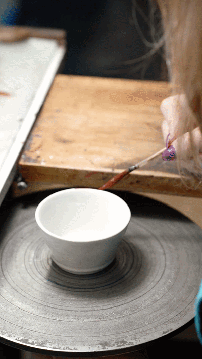 Hands hand-painting the red rim of a white Beja bowl on a potter's wheel