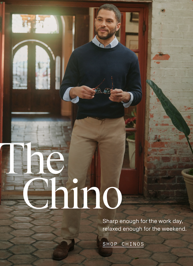 Man standing outdoors on a tiled patio in front of an arched wooden door, wearing khaki chinos, a navy crewneck sweater layered over a light blue dress shirt, and dark brown loafers, holding sunglasses. Text reads: "The Chino. Sharp enough for the work day, relaxed enough for the weekend. SHOP CHINOS"