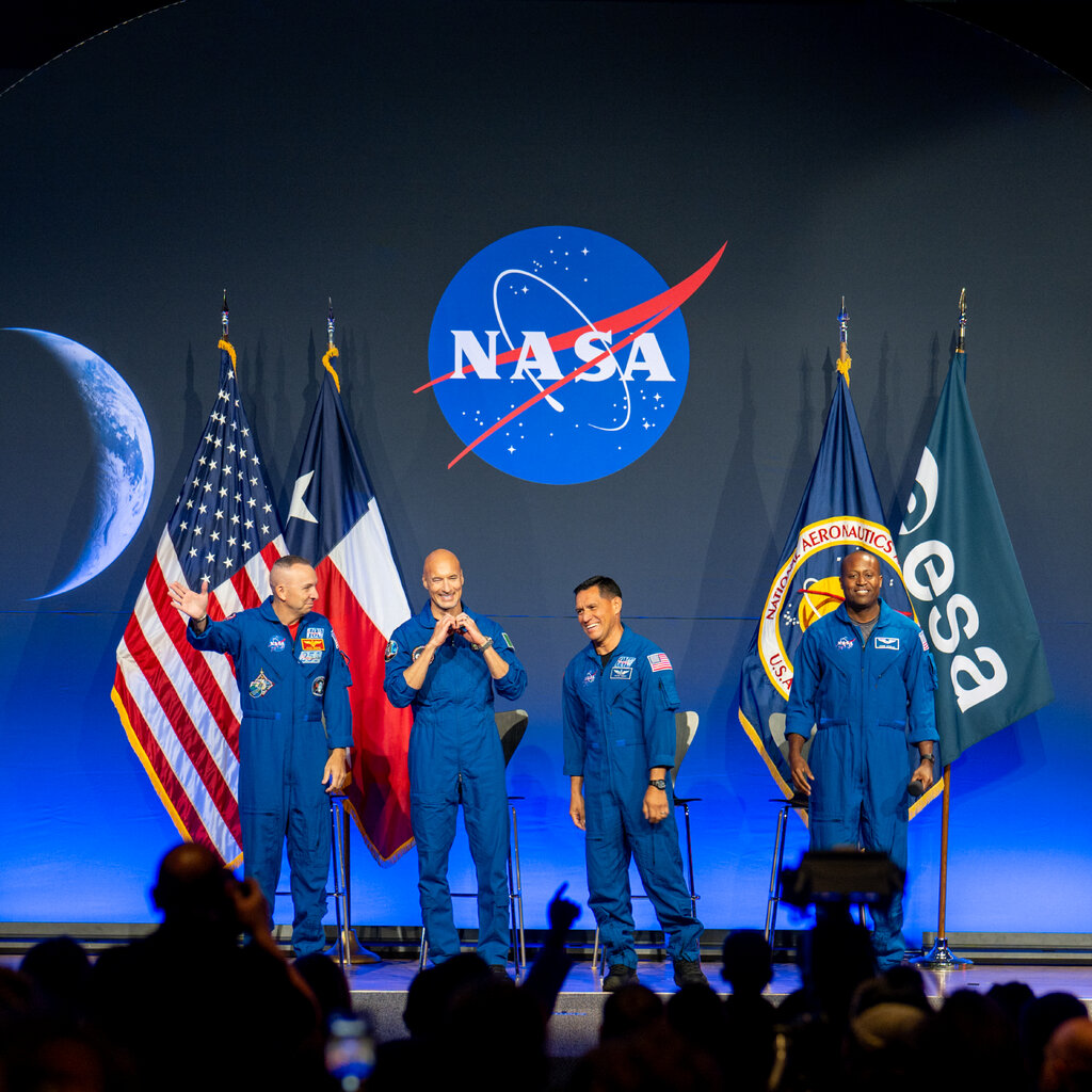 From left, Randy Bresnik, Luca Parmitano, Frank Rubio and Andre Douglas are onstage wearing blue jumpsuits. Behind them are flags and a large NASA logo.
