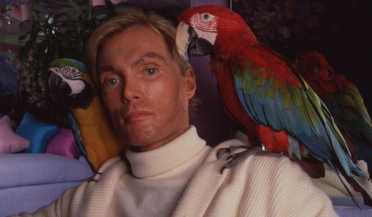 A man poses with two parrots on his shoulders.