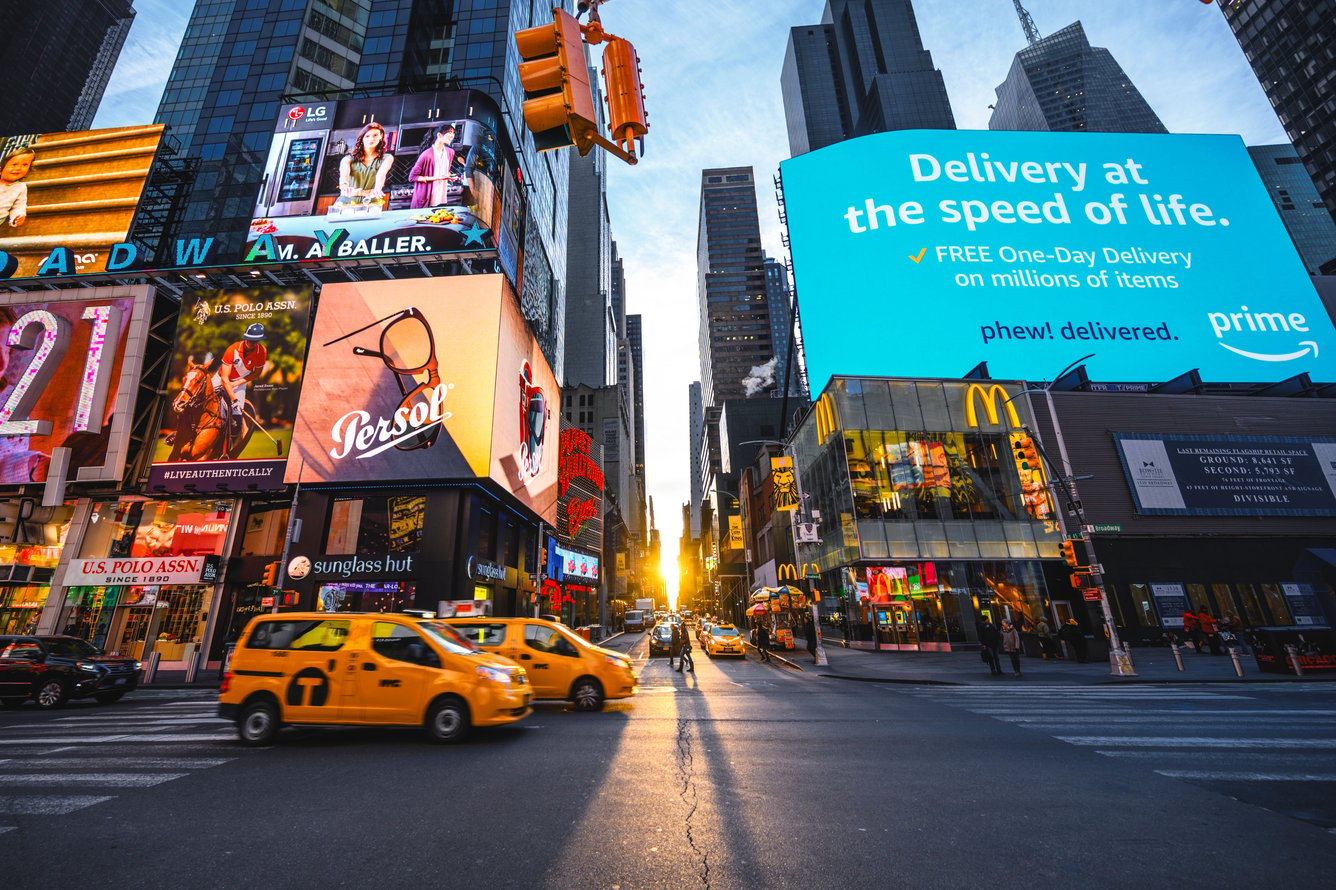 digital billboards screens are shown at a corner of Times Square as the sun sets, with taxis crossing the street in front of the screens