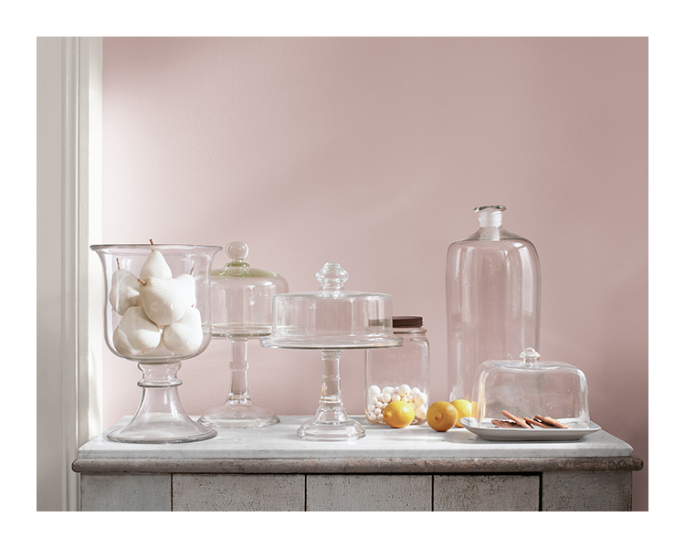 A gray, weathered accent table is topped with glass cake stands and cloches of various heights in front of a pale purple wall.