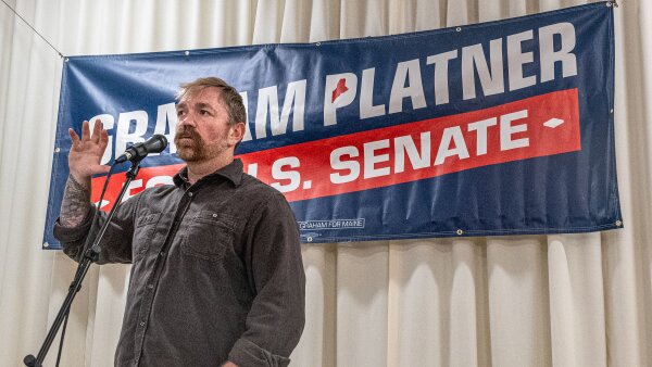 Graham Platner speaks to voters at a town hall. He wears a casual dark button-up shirt. 