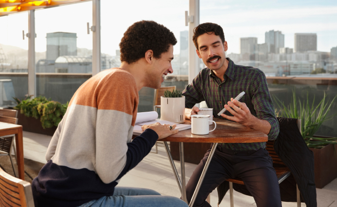 Two men sit at a rooftop patio table looking at real estate on the Zillow app.