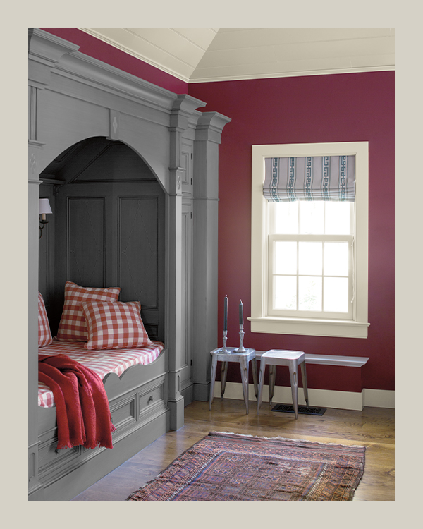 A plum-painted bedroom with wood floors, a decorative area rug, and a gray-painted built-in bed with red checkered bedding.