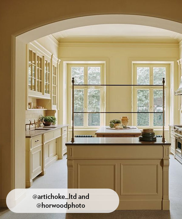 A soft, buttery-yellow kitchen with classic cabinetry, tall windows, and a central island framed by an arched wall. Natural light floods the room, creating a serene atmosphere enhanced by simple styling and warm traditional details.