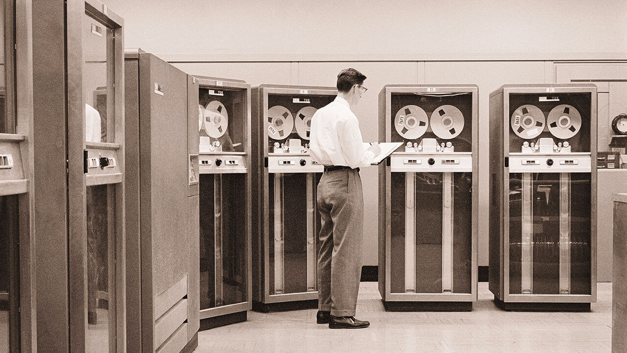 A computer technician stands before an array of ibm model 727 magnetic tape data storage drive units.