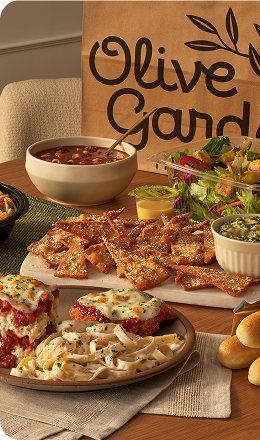 Olive Garden meal spread with pasta, salad, soup, breadsticks, and appetizer bites in front of a branded takeout bag.