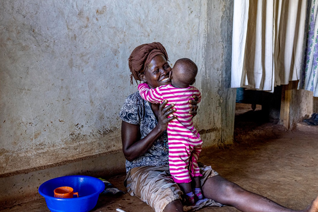 A woman sitting on the floor, holding a baby in a striped onesie, with a warm smile on her face