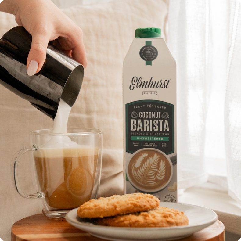 A person pouring frothed Elmhurst Coconut Barista milk into a glass latte cup next to a plate of cookies