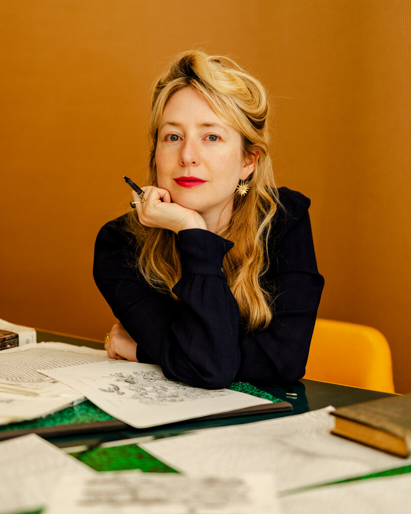 Joana Avillez sits at a table covered in drawings, holding a pen and looking into the camera lens.
