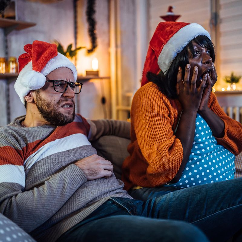 Two figures wearing Santa hats sit on a couch gasping at a TV screen.