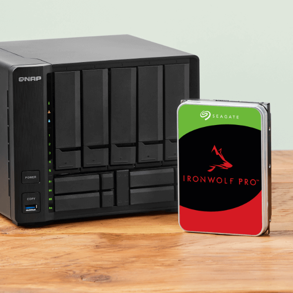 A black QNAP NAS device with multiple drive bays sits on a wooden table next to a Seagate IronWolf Pro hard drive.
