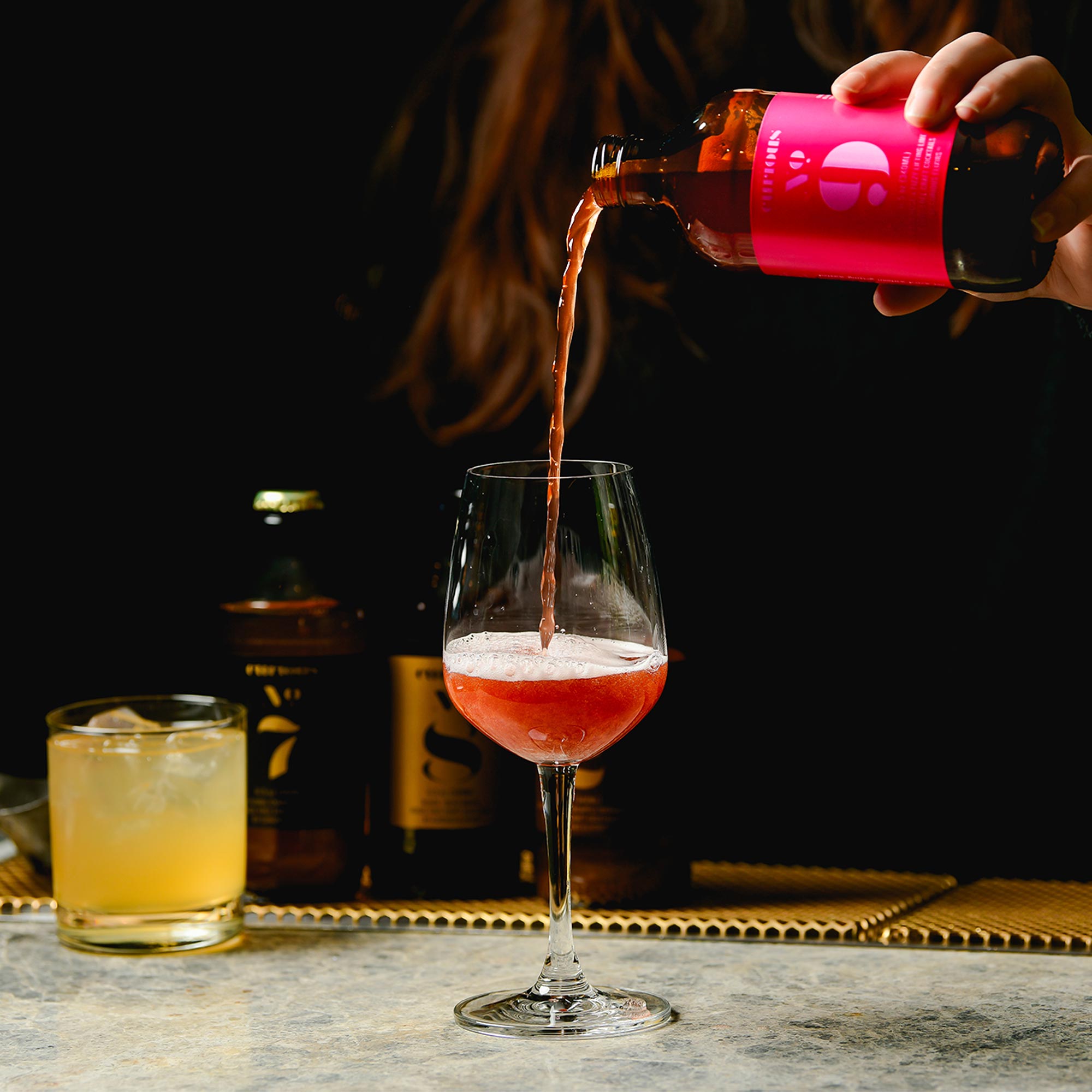 Someone pours a bottle of Curious No. 9 into a wine glass. In the background are bottles and glasses of Curious No. 7 and No. 8.